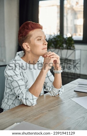 thoughtful non-binary manager with hands near chin sitting at workplace in office and looking away
