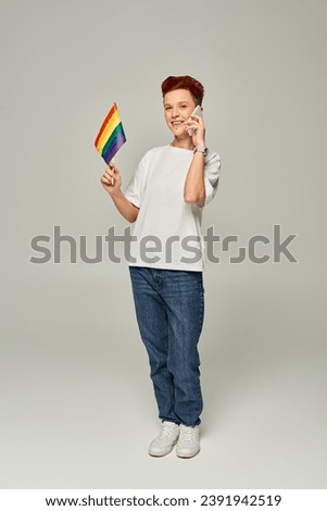 happy redhead queer person holding small LGBT flag and talking on smartphone on grey, banner