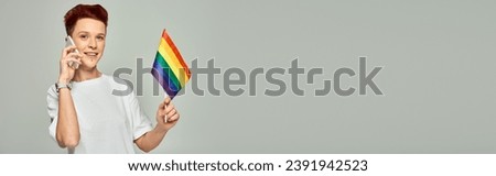 happy redhead queer person holding small LGBT flag and talking on smartphone on grey, banner