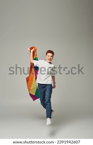 overjoyed queer person in white t-shirt and jeans and levitating with LGBT flag on grey backdrop