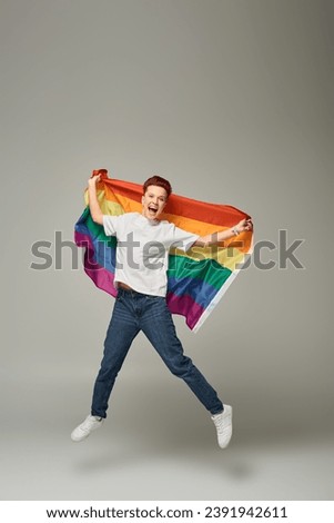 overjoyed queer person in white t-shirt and jeans jumping and levitating with LGBT flag on grey