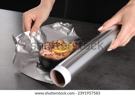 Woman wrapping tasty salmon with lemon and spices in aluminum foil at grey textured table, closeup Royalty-Free Stock Photo #2391957583