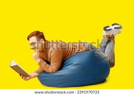 Young man lying on beanbag and reading book on yellow background