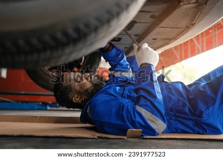 Mechanic in blue uniform lying down and working under car at auto service garage. Portrait of a happy mechanic man working on a car in an auto repair shop.