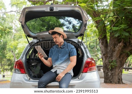 Asian man sitting back of car and holding tables check location and find place to travel. Happy road trip and vacation in summer in the countryside. Concept of journey on leisure, road trip vacation
