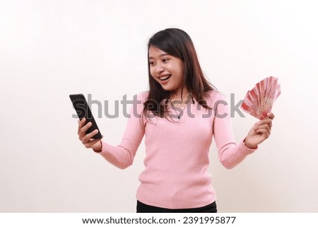 Surprised young asian girl getting good news from her phone while holding Indonesia banknotes. Isolated on white
