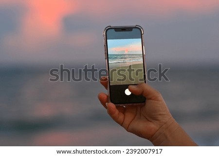 Close-up of an old woman's hand holding a phone to take a photo of a beautiful tropical beach in the evening.