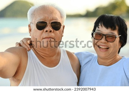 Portrait of Senior Asian couple looking to the camera taking a selfie together and enjoying at the beach, Two active seniors traveling outdoors concept.