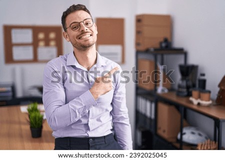 Young hispanic man at the office cheerful with a smile on face pointing with hand and finger up to the side with happy and natural expression 