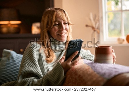 Woman At Home With Warming Hot Drink Of Tea Or Coffee In Cup Or Mug With Mobile Phone Streaming Royalty-Free Stock Photo #2392027467