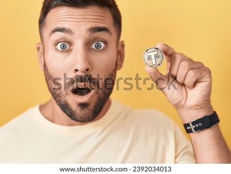 Handsome hispanic man holding tether cryptocurrency coin scared and amazed with open mouth for surprise, disbelief face  Royalty-Free Stock Photo #2392034013