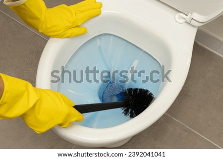 Close up of hands wearing yellow gloves cleaning a modern, white, ceramic toilet bowl with a black toilet brush and using blue liquid toilet cleaner
