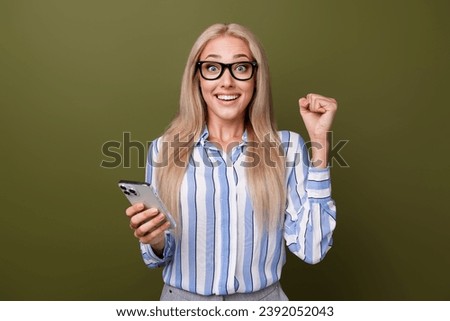 Photo portrait of blonde lovely young lady hold device excited raise fists wear trendy striped garment isolated on khaki color background