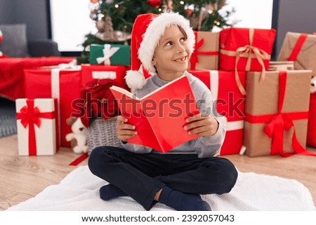 Adorable hispanic boy reading book sitting on floor by christmas tree at home
