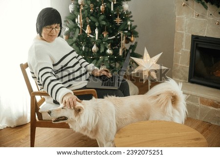 Happy senior woman using laptop and playing with adorable white dog in stylish festive christmas living room. Beautiful mature woman freelancer having remote work at home with pet at winter holiday