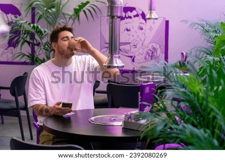 a young guy is sitting at a table in a cafe restaurant in a beautiful interior waiting for his order with smartphone in his hand