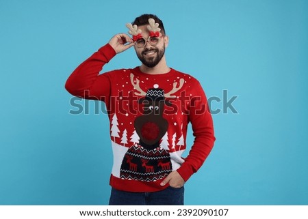 Happy young man in Christmas sweater and funny glasses on light blue background
