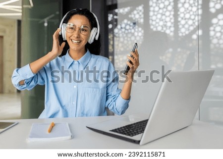 Young beautiful woman at workplace joyful listening to music and online radio, using app on phone, businesswoman resting on break, singing along well done job well done. Royalty-Free Stock Photo #2392115871