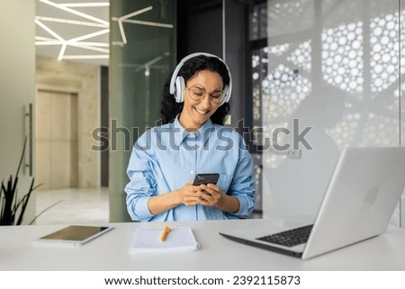 Young beautiful woman at workplace joyful listening to music and online radio, using app on phone, businesswoman resting on break, singing along well done job well done. Royalty-Free Stock Photo #2392115873