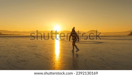 LENS FLARE, SILHOUETTE: Early winter morning with lady skating on a scenic pond. She is ice skating while first golden rays of sun peek over hills and bounce off the surface of a naturally frozen lake