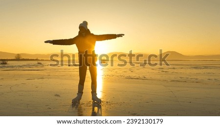 LENS FLARE, SILHOUETTE: A happy woman is skating on a golden shining frozen lake. She is having fun and enjoys dancing on vast empty natural ice rink as the winter sun is setting behind the hills.