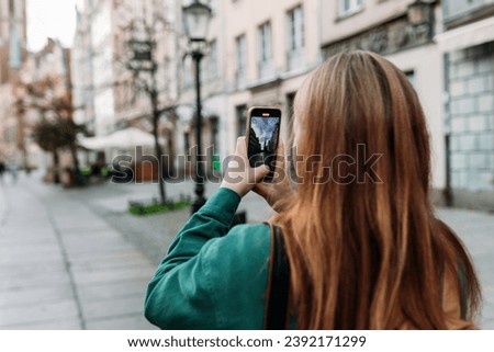 Person in fashion clothes is recording content with a smartphone on urban background. Stylish 30s girl with phone walking outdoors
