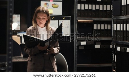 Female private detective conducting investigation in office, focused on reviewing files and evidence. Portrait shot of policewoman analyzing statements and records. Looking at camera, tripod shot. Royalty-Free Stock Photo #2392175261