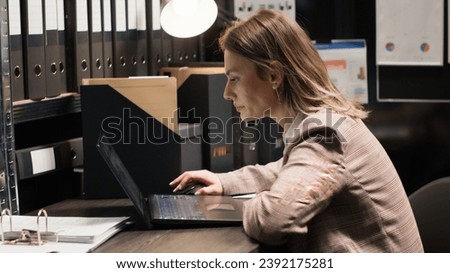Policewoman captured in midst of investigation, examining records and evidence. Detailed view of officer at desk with laptop and case files opened for cross-referencing suspect profile and statements. Royalty-Free Stock Photo #2392175281