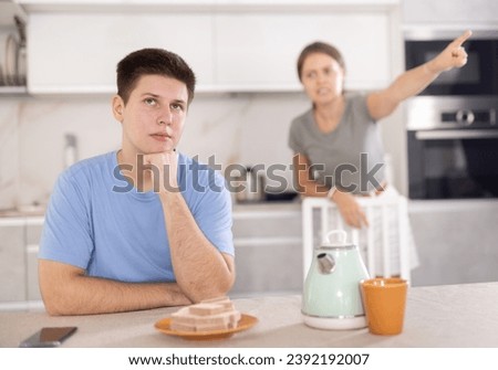 Frustrated offended guy sitting at kitchen table on background of swearing dissatisfied young wife. Family conflicts and communication problems concept Royalty-Free Stock Photo #2392192007