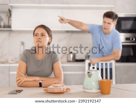 Sad young girl sitting at the table while her boyfriend is quarreling with her standing behind in the kitchen Royalty-Free Stock Photo #2392192147