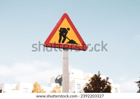 Roadworks yellow red triangular sign on a city street against blue sky. Temporary construction icon in urban setting. Work in progress. Man is working hard outdoors signboard. Urban landscape.