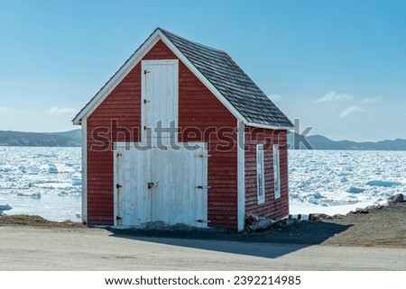 An old two story vibrant red barn with double white doors and a hay loft. The roof is made of black cedar shakes. The shed is at the edge of an icy shore. The harbor is filled with thick slob ice.