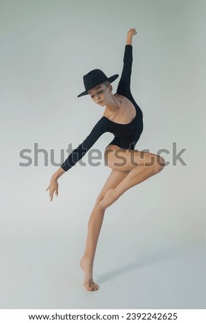 a young ballerina in a black bodysuit shows ballet steps in motion standing on one leg and spreading her hands in a hat