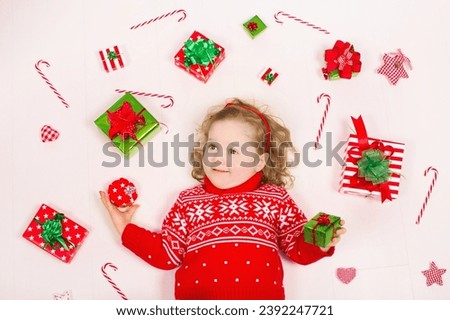 Child opening Christmas presents. Little girl in knitted winter sweater with present box. Kids open gifts. Toddler kid on the floor under decorated Xmas tree. Children play with gift box and candy.