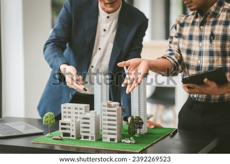 Two middle-aged Asian people businessmen, one holding wind turbine model, discussing over model city, likely about clean energy and new housing projects. Royalty-Free Stock Photo #2392269523