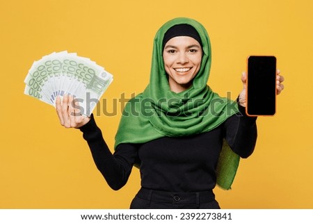 Young muslim woman wear green hijab abaya black clothes hold fan of dollar cash money use blank screen mobile cell phone isolated on plain yellow background. People uae middle eastern islam concept