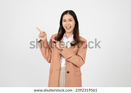 Young asian business woman, smiling while pointing finger left and showing thumbs up, recommending product, praise, standing over white background. Royalty-Free Stock Photo #2392319561