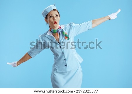 happy stylish female stewardess on blue background in blue uniform with outstretched arms imitating flight of airplane. Royalty-Free Stock Photo #2392325597