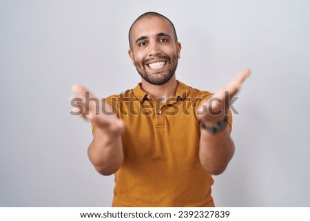 Hispanic man with beard standing over white background smiling cheerful offering hands giving assistance and acceptance. 