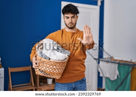Hispanic man with beard holding laundry basket at laundry room with open hand doing stop sign with serious and confident expression, defense gesture 
