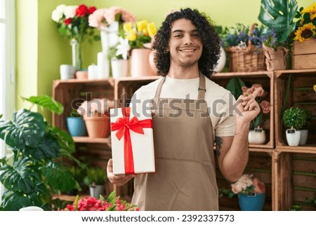 Hispanic man with curly hair working at florist shop holding gift smiling happy pointing with hand and finger to the side 