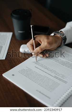 A person wearing a white long-sleeved shirt, sitting at a wooden desk, where they are in the process of signing a document with a silver pen