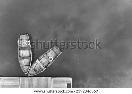 An artistic, monochrome, perpendicular view of two wooden boats at a pier on the blue-green surface of Lago di Braies in the Dolomites. Ideal for black and white poster projects.