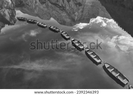Monochrome, artistic, perpendicular view of wooden boats in row among mirroring peaks and sky on a lake Lago di Braies in the Dolomites. Ideal for black and white poster projects.
