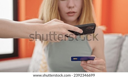 Young blonde woman make photo to credit card by smartphone sitting on sofa at home