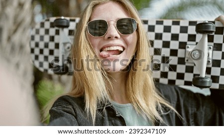 Young blonde woman holding skate make selfie by camera with tongue out at park