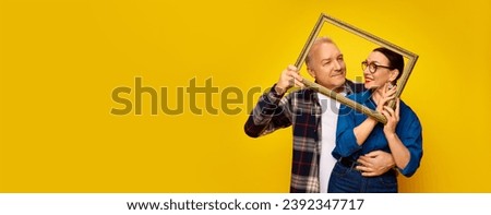Portrait of happy, loving, senior couple, man and woman posing with picture frame against yellow studio background. Concept of marriage, relationship, Valentine's Day, love, emotions. Banner