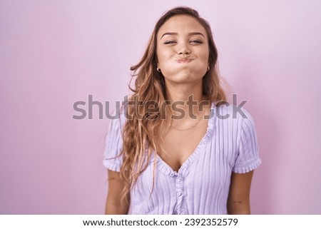 Young hispanic woman standing over pink background puffing cheeks with funny face. mouth inflated with air, crazy expression.  Royalty-Free Stock Photo #2392352579