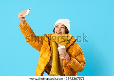 Young Asian woman in winter clothes with coffee cup taking selfie on blue background