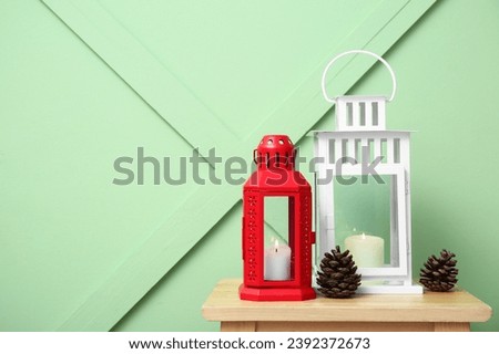 Christmas lanterns with burning candles and pine cones on wooden stool near color wall
